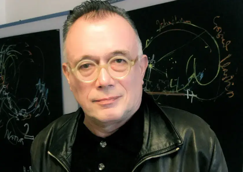 Un homme portant des lunettes rondes et une veste en cuir, se tenant devant un tableau noir avec des dessins abstraits.
