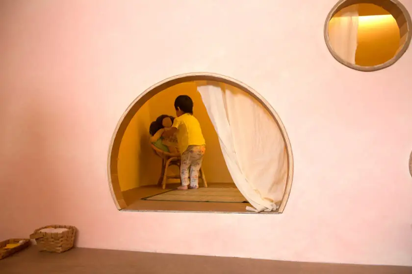 Un enfant joue dans une alcôve ronde d'une pièce aux murs roses, avec un autre enfant assis sur une chaise.