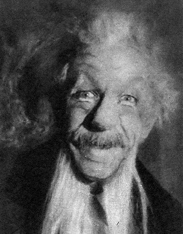 Un homme âgé avec une grande moustache et des cheveux ébouriffés, souriant à la caméra.