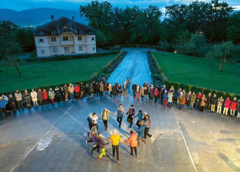 Un groupe de personnes danse en cercle sur une allée, entouré d'arbres et d'un bâtiment en arrière-plan.