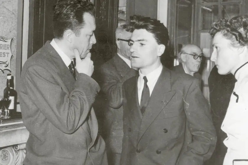 Deux hommes en costume discutent dans un cadre intérieur, avec une femme à droite et d'autres personnes en arrière-plan.