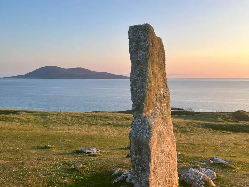 Un menhir en pierre sur un terrain verdoyant, face à la mer et une colline au loin, sous un ciel au crépuscule.