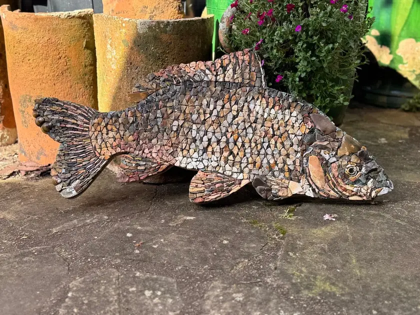 Sculpture en mosaïque représentant un poisson, posée sur un sol en pierre avec des pots de fleurs en arrière-plan.