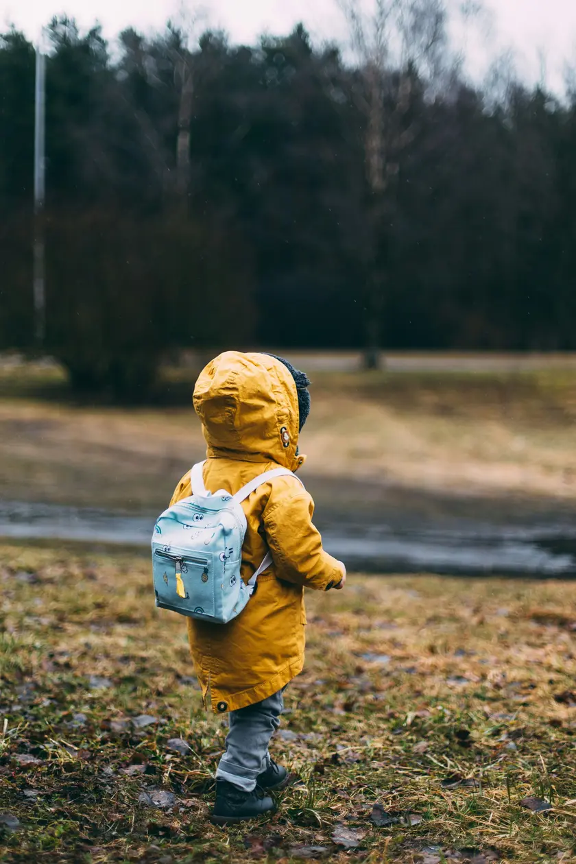 Un enfant de dos portant un manteau jaune et un petit sac à dos bleu, se tenant sur un terrain herbeux.
